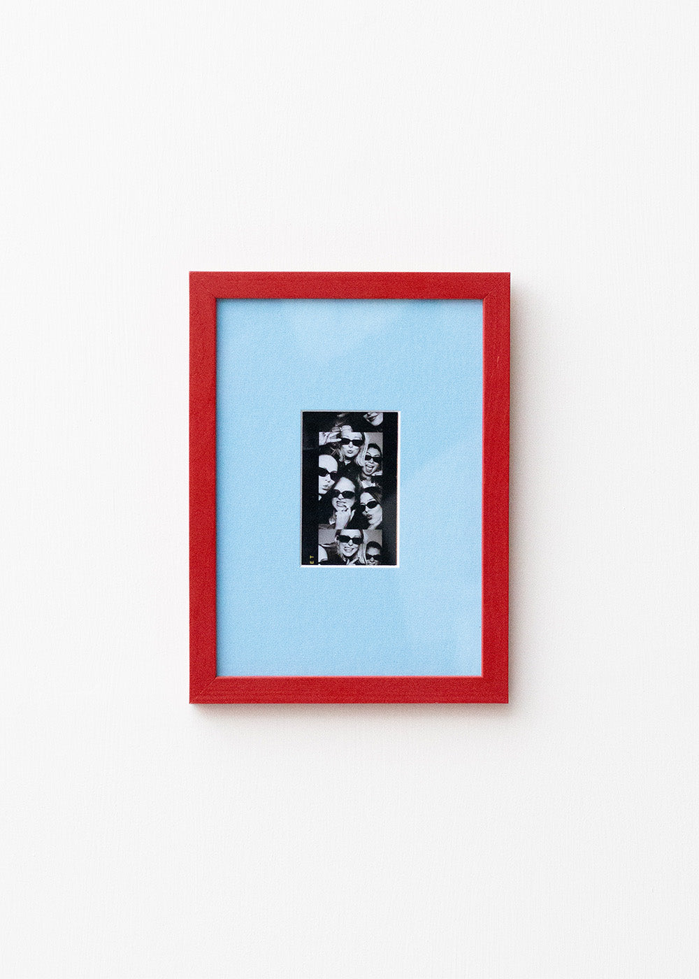 A framed art print featuring a black and white collage in a vertical black frame, set against a light blue mat and a vibrant red outer frame, mounted on a plain white wall.