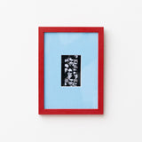 A framed art print featuring a black and white collage in a vertical black frame, set against a light blue mat and a vibrant red outer frame, mounted on a plain white wall.