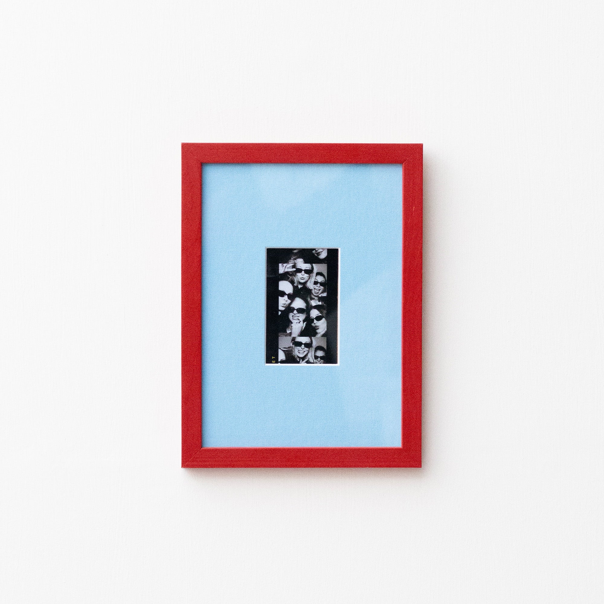 A framed art print featuring a black and white collage in a vertical black frame, set against a light blue mat and a vibrant red outer frame, mounted on a plain white wall.