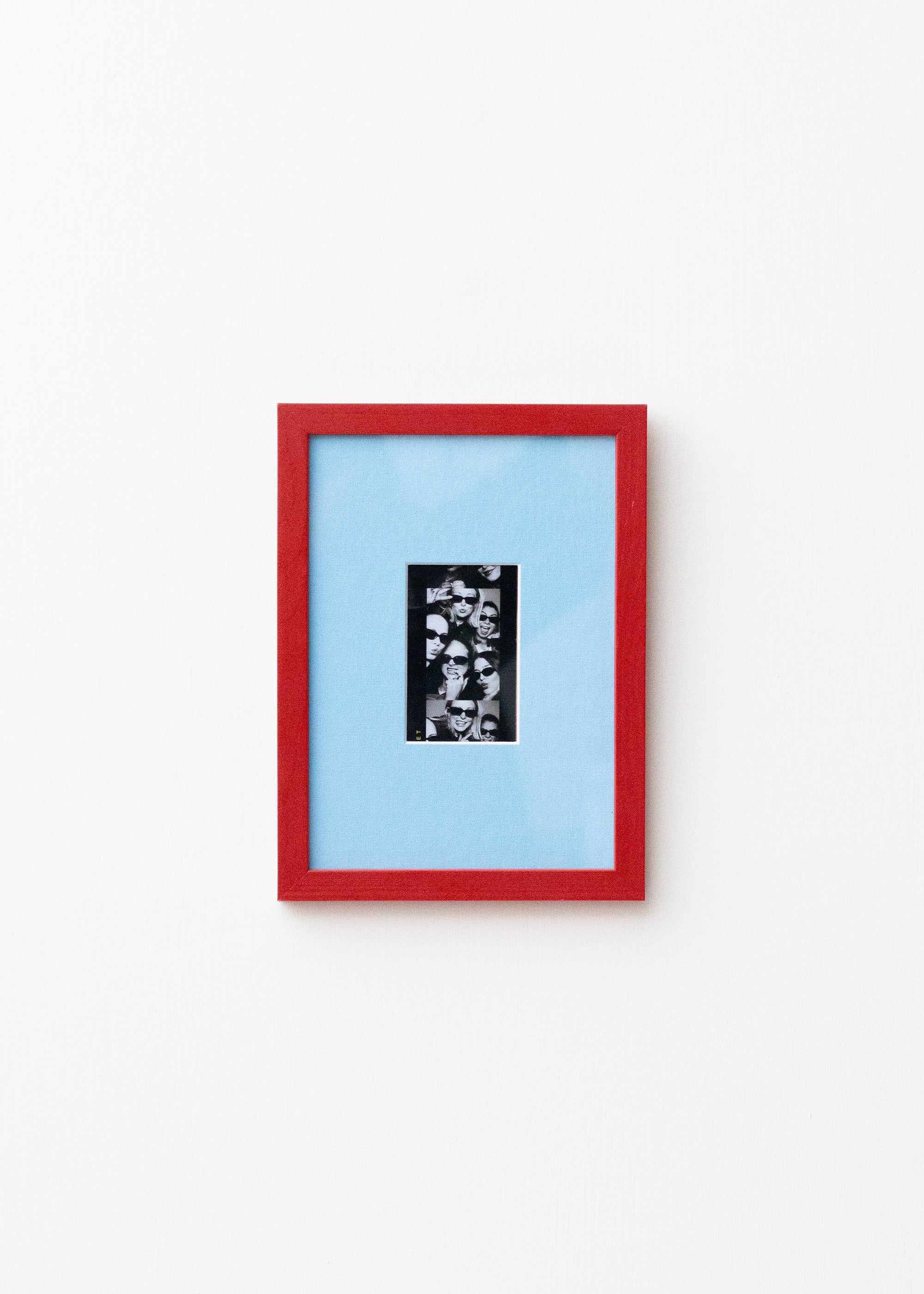 A framed art print featuring a black and white collage in a vertical black frame, set against a light blue mat and a vibrant red outer frame, mounted on a plain white wall.