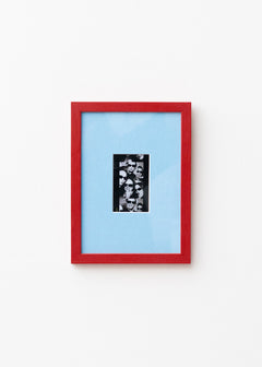 A framed art print featuring a black and white collage in a vertical black frame, set against a light blue mat and a vibrant red outer frame, mounted on a plain white wall.