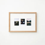 A light natural wood frame with a white mat holding three small landscape photographs, displayed against a plain white wall.