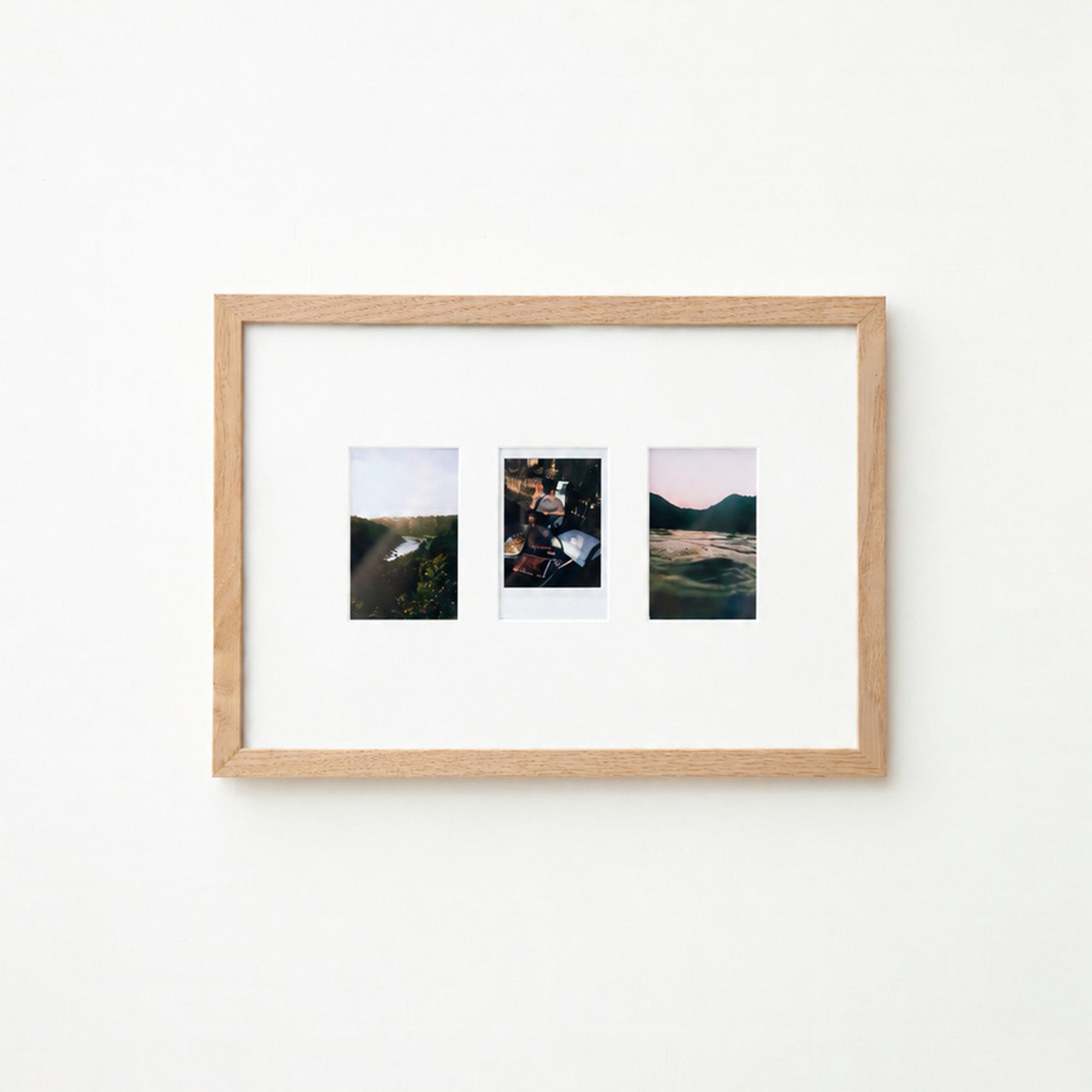 A light natural wood frame with a white mat holding three small landscape photographs, displayed against a plain white wall.