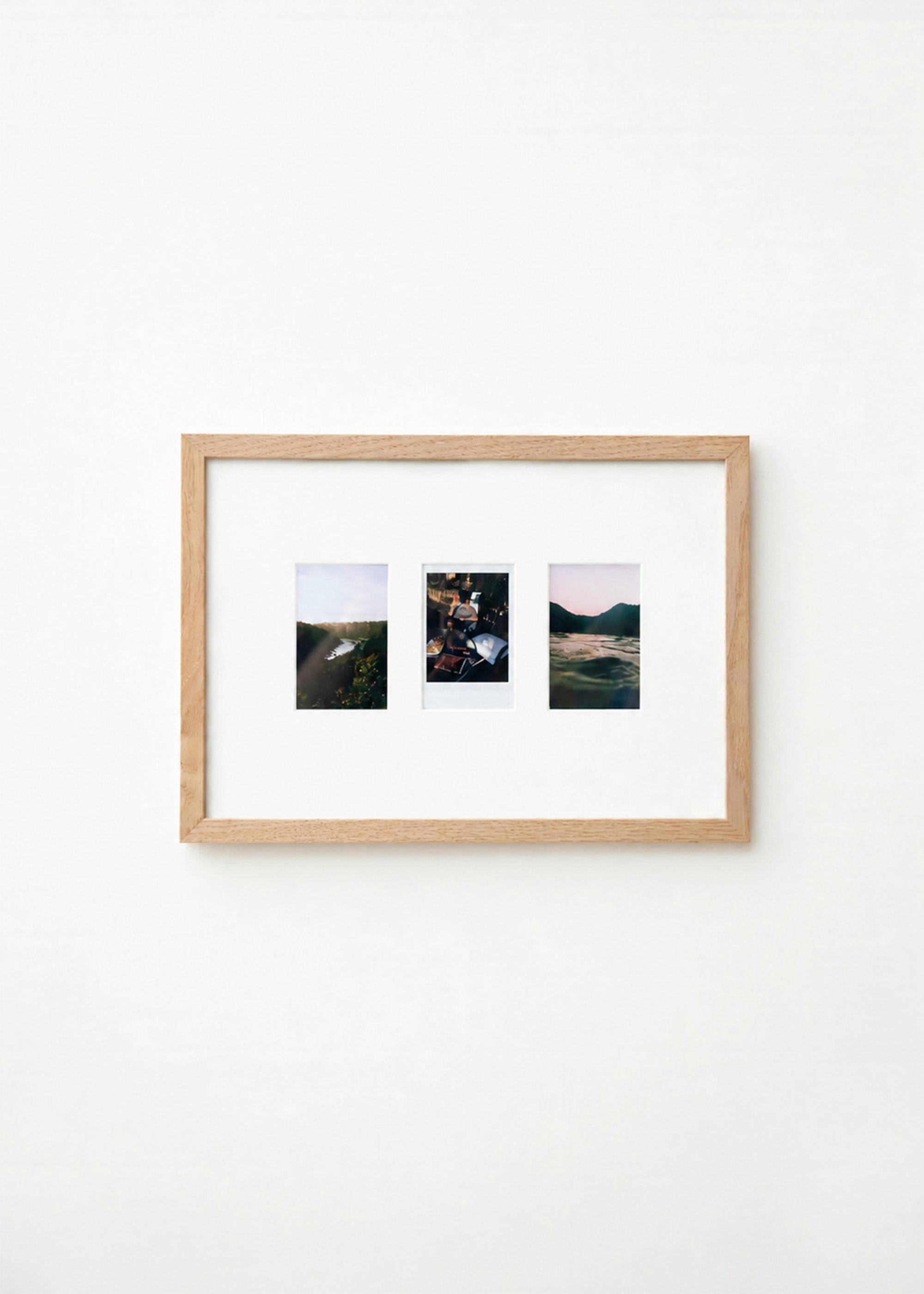 A light natural wood frame with a white mat holding three small landscape photographs, displayed against a plain white wall.