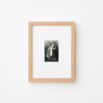 A small black and white portrait photograph in a light natural wood frame with a white mat, mounted on a plain white wall.