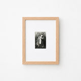 A small black and white portrait photograph in a light natural wood frame with a white mat, mounted on a plain white wall.