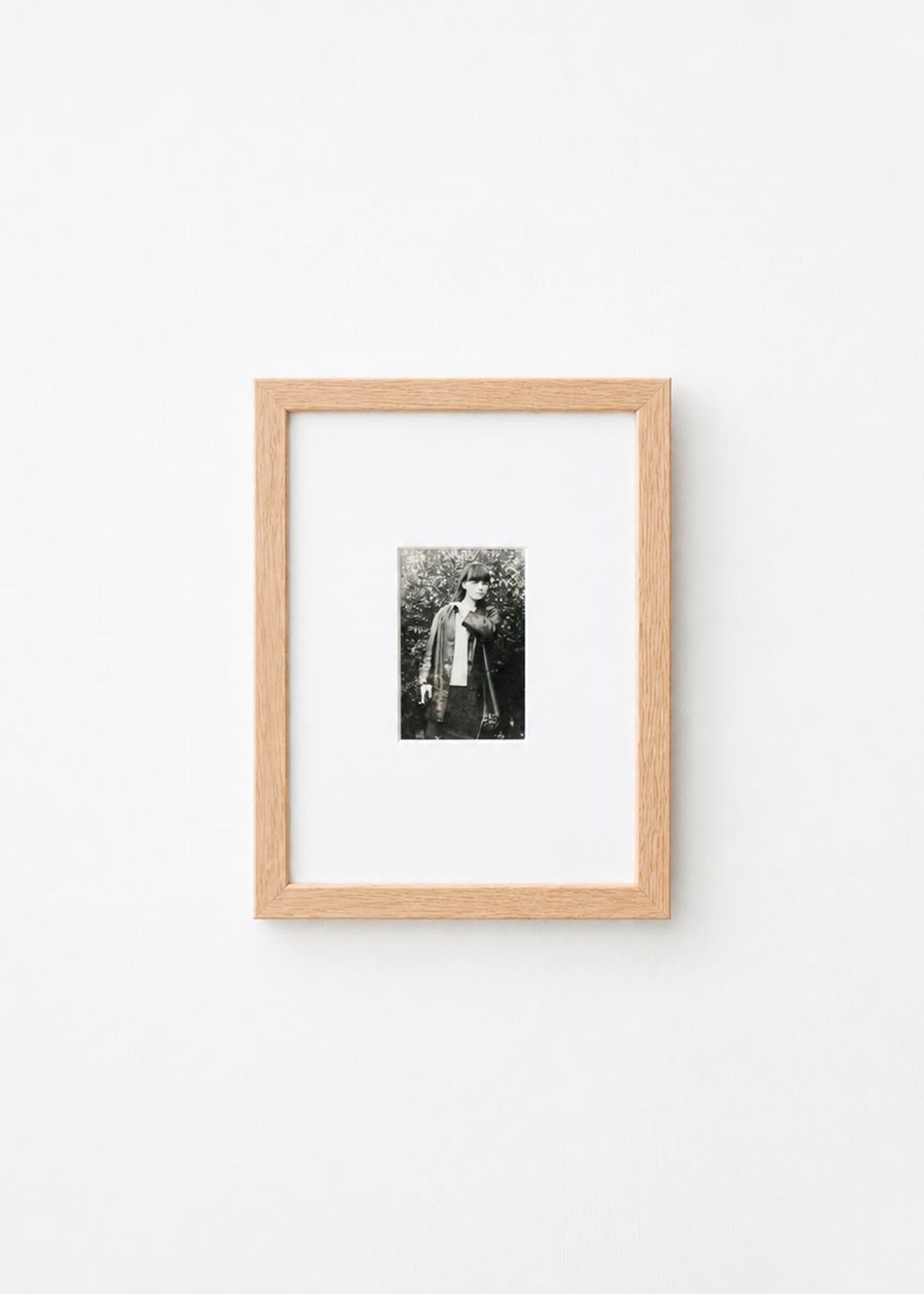 A small black and white portrait photograph in a light natural wood frame with a white mat, mounted on a plain white wall.