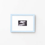 A square light blue picture frame with a white mat, containing a small black-and-white photograph, shown hanging on a plain white wall.