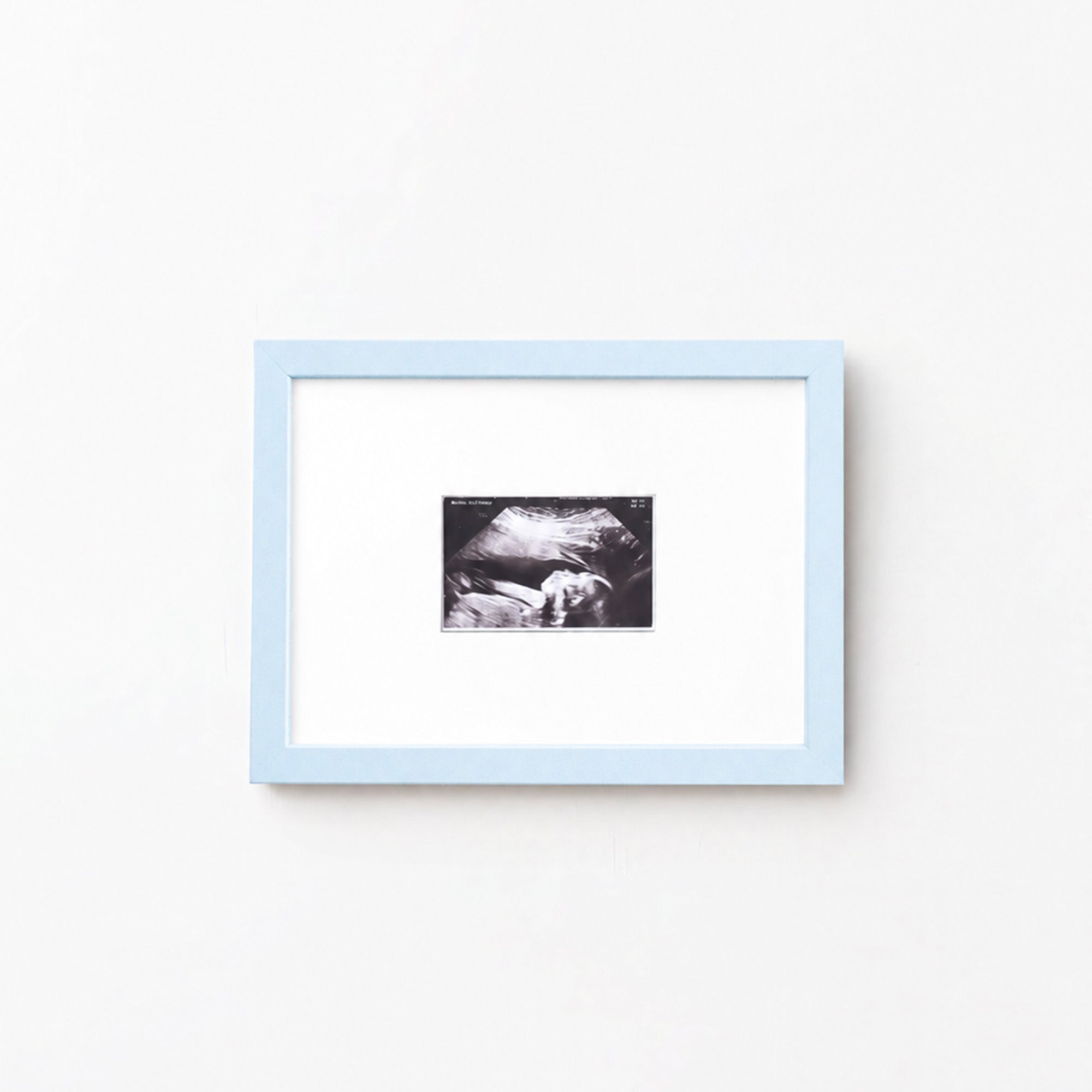 A square light blue picture frame with a white mat, containing a small black-and-white photograph, shown hanging on a plain white wall.