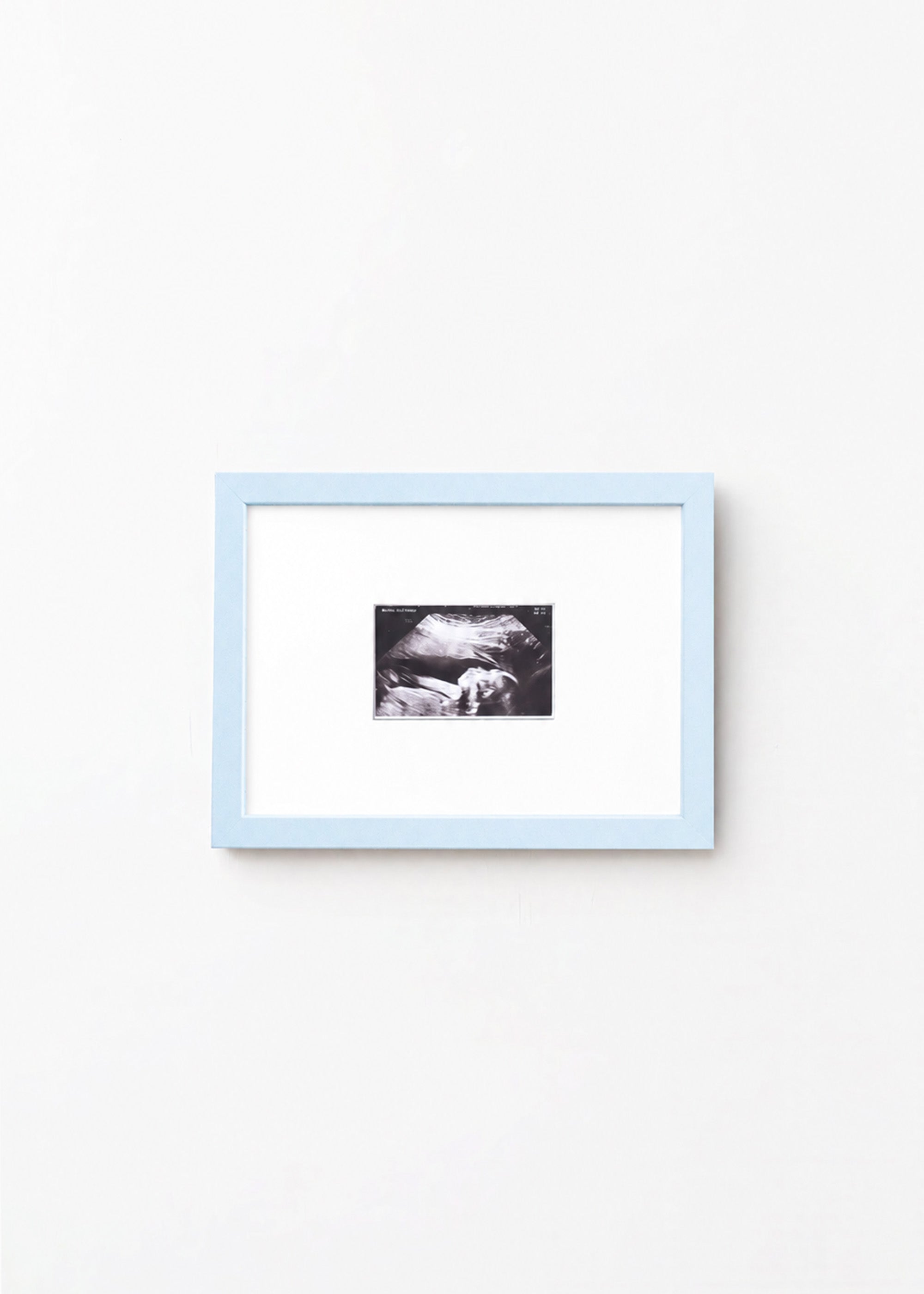 A square light blue picture frame with a white mat, containing a small black-and-white photograph, shown hanging on a plain white wall.