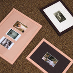 Three framed picture frames arranged on a brown carpeted floor. Two frames have light pink borders with white mats and small photos, and one frame has a dark brown border with a white mat and a larger photo.