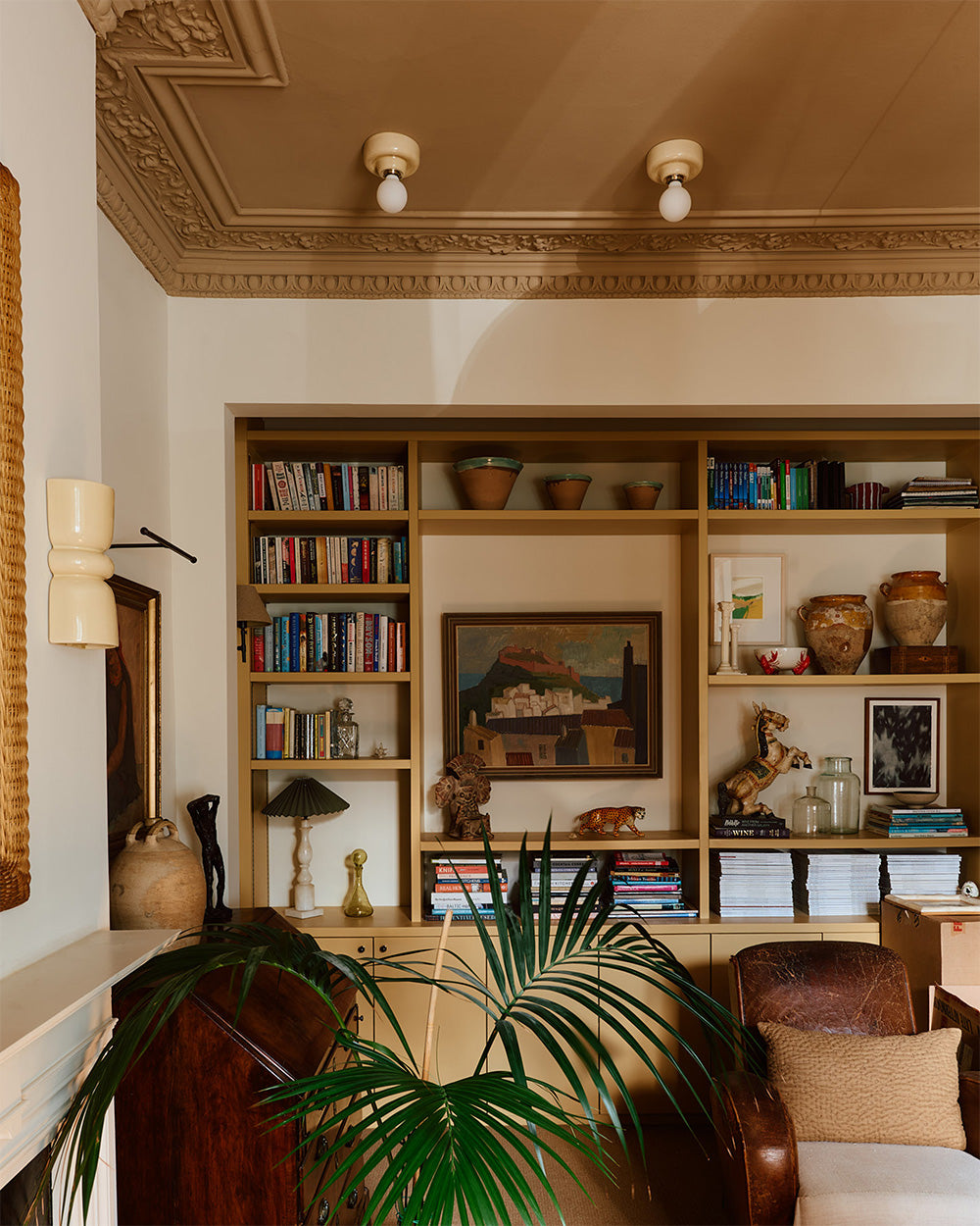 A warm, styled interior corner featuring a large built-in wooden bookshelf filled with books, ceramic vases, and framed art. A green palm plant is in the foreground, with a brown leather armchair and a cream wall visible.