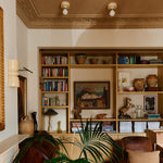 A warm, styled interior corner featuring a large built-in wooden bookshelf filled with books, ceramic vases, and framed art. A green palm plant is in the foreground, with a brown leather armchair and a cream wall visible.