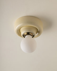 A classic ceiling-mounted light fixture with a round butter yellow-coloured ceramic or plaster canopy and a frosted white globe bulb, shown against a matching textured off-white ceiling.