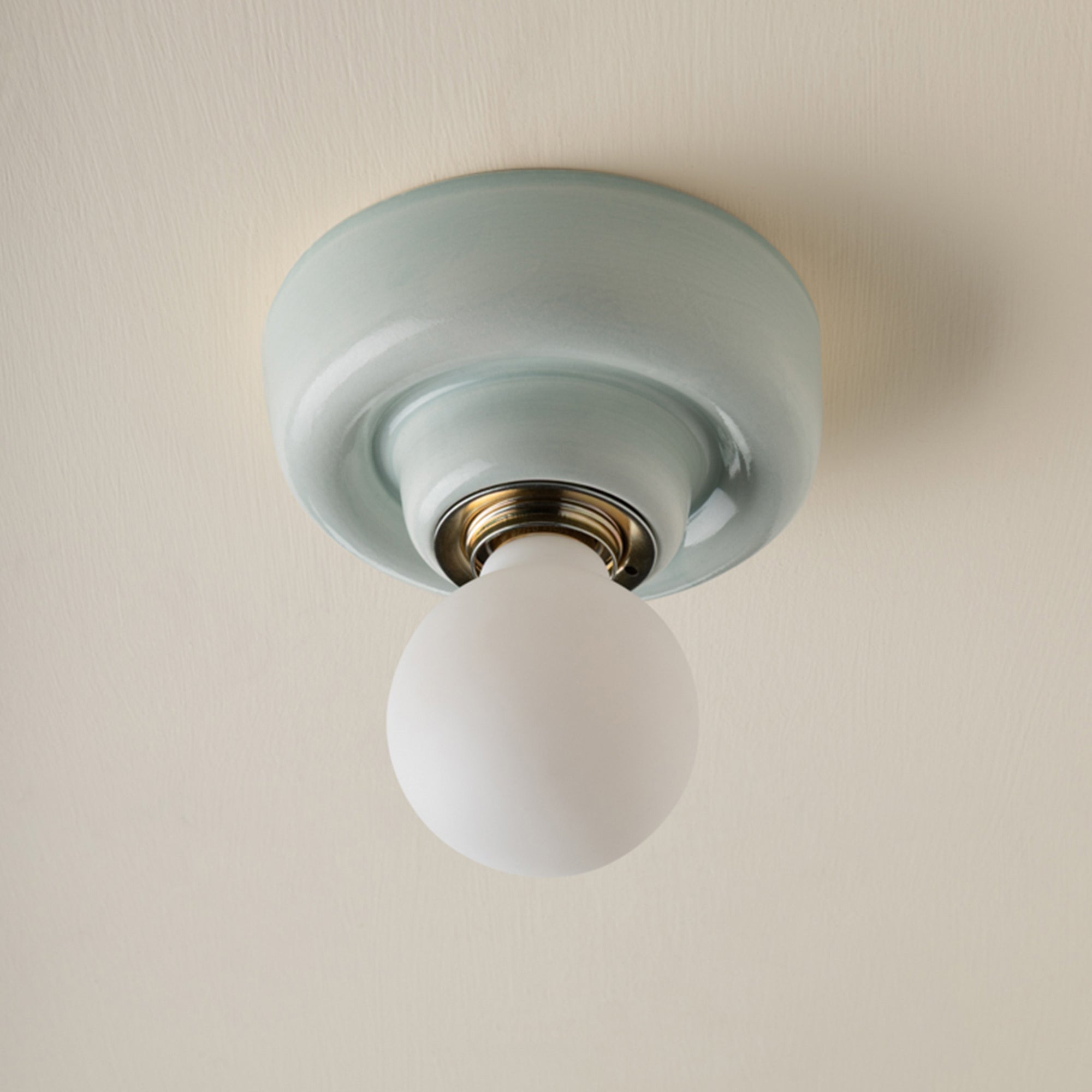 A ceramic ceiling-mounted light fixture in pale blue-green and a frosted white globe bulb, shown against a warm off-white ceiling.