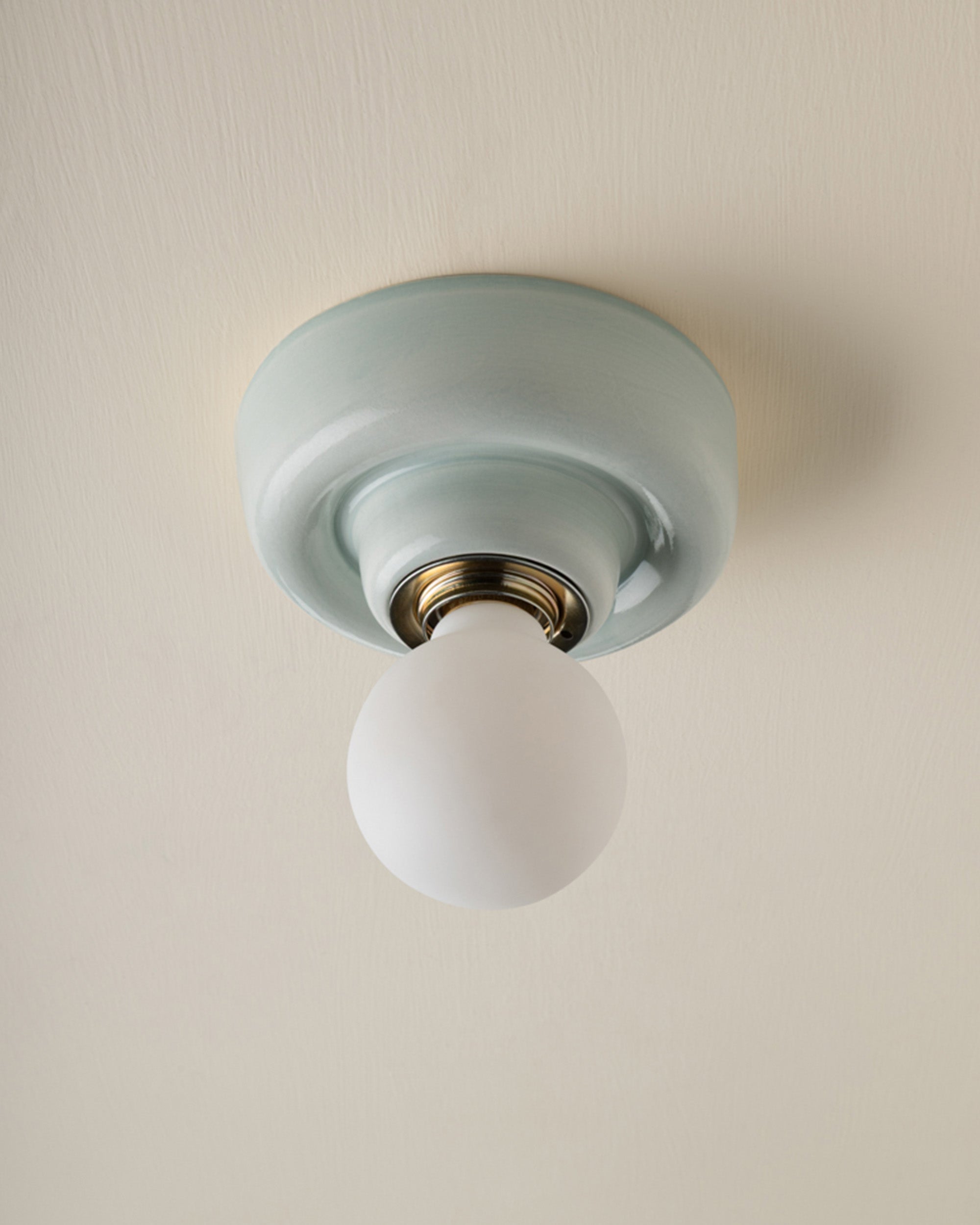 A ceramic ceiling-mounted light fixture in pale blue-green and a frosted white globe bulb, shown against a warm off-white ceiling.