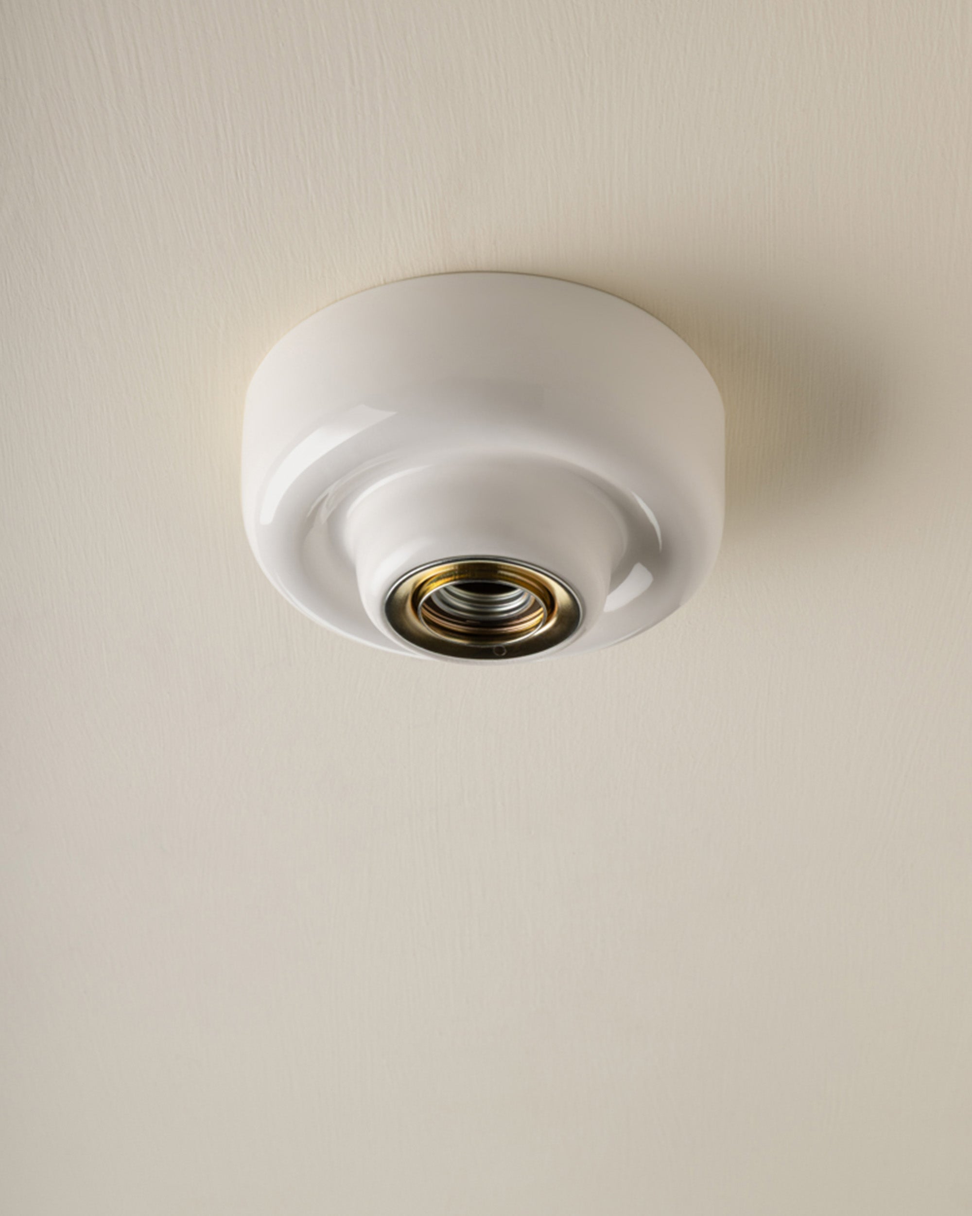 A white ceramic flush-mount ceiling light fixture with a gold-toned metal socket, shown installed on a textured off-white ceiling.