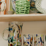 Colorful glassware on a shelf with a green woven basket, glassware and abstract painting in the background.