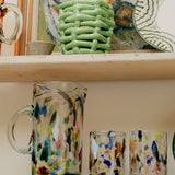 Colorful glassware on a shelf with a green woven basket, glassware and abstract painting in the background.
