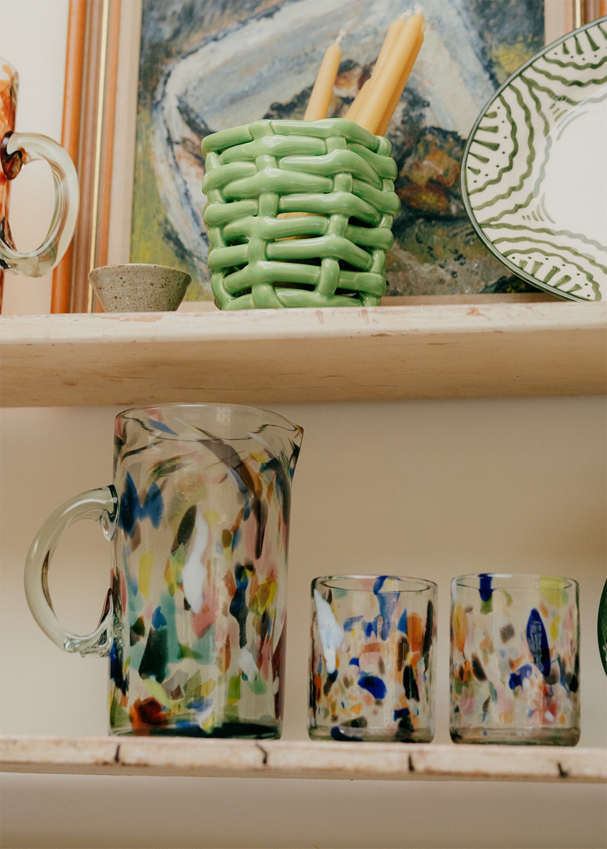 Colorful glassware on a shelf with a green woven basket, glassware and abstract painting in the background.