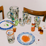 Cocktsil table with colorful glassware, plates, and a knife on a white tablecloth.